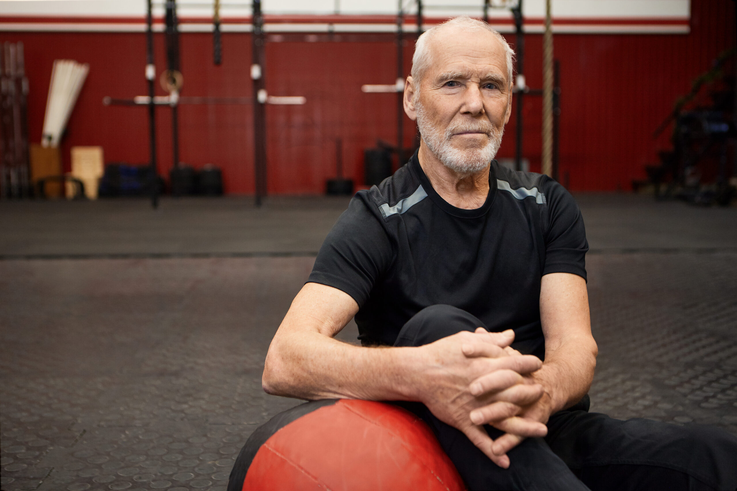 Confident elderly man with a beard sitting on the gym floor, resting after a workout with a red exercise ball in a modern fitness facility.